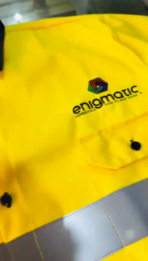 Close view of a yellow work shirt showing the embroidered Enigmatic logo, a buttoned chest pocket and the stitching on a grey reflective band