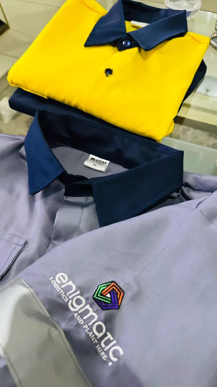 A folded yellow and navy work shirt stacked on a grey work shirt with an embroidered Enigmatic Logistics and Plant Hire logo on the sleeve, made by BMC Manufacturing