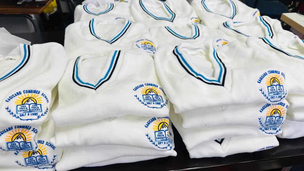 Stacks of white knitted school jerseys with blue and black V-neck trim, each with an embroidered Kagisano Combined School crest